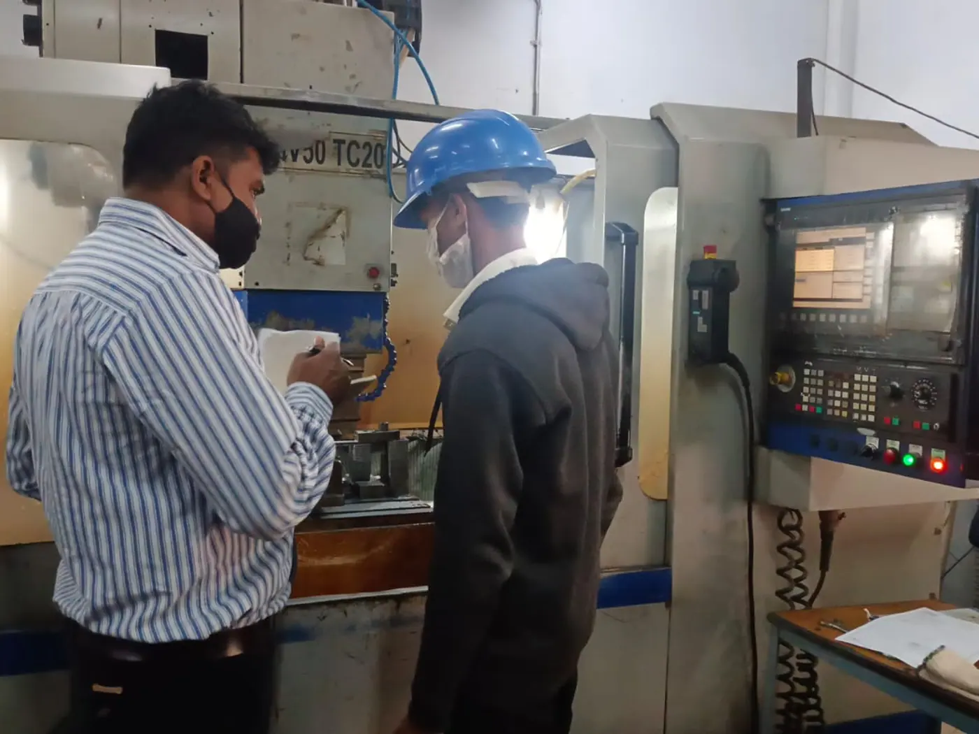 A trainer and a trainee in a hard hat talking through a job at a CNC machine